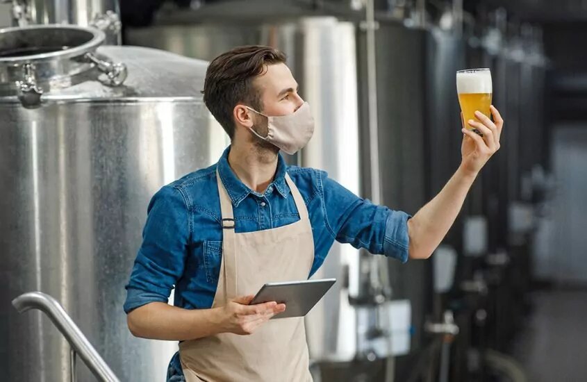 Brewer is a face mask holding up a glass of beer while inspecting it