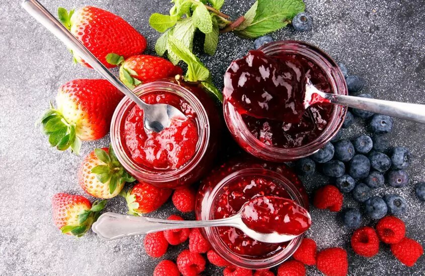 Jar of Strawberry jam next to a jar of raspberry jam next to a jar of blueberry jam all surrounded by their respective fruits