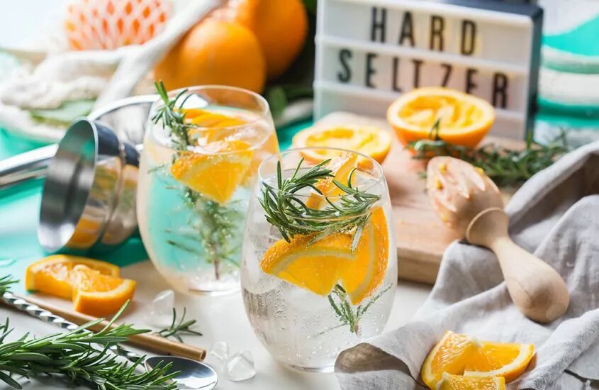 Two glasses of hard seltzer with pieces of orange in the surrounding area and a sign in the background that says "Hard Seltzer"