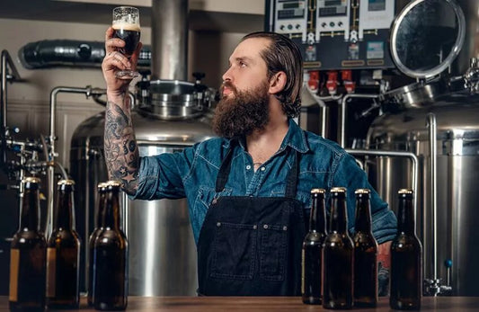 A man in a brewery is holding a glass of beer up while inspecting it. 
